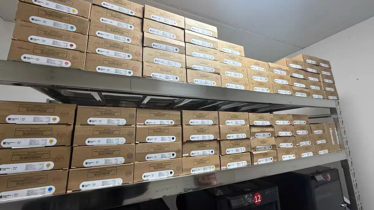 Filament Storage Ideas: 6 Setups Ranked by Cost, Capacity, and What They Don't Fix 2 Two shelves of boxed PLA filament stacked above a row of 3D printers in a workshop