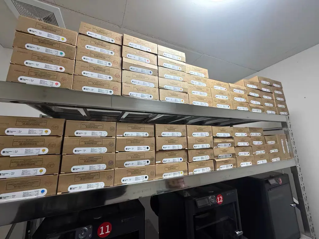 Heat Resistant 3D Printing Filament: What Actually Holds Up (And What the Charts Get Wrong) 4 Shelves stacked with boxed PLA filament above a row of running 3D printers