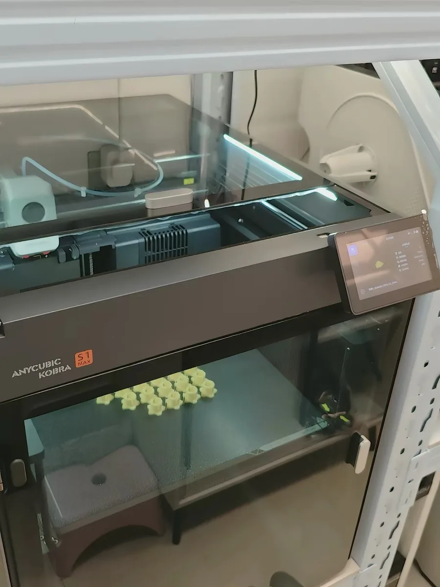 The orange S1 MAX badge on the front of the Anycubic Kobra S1 Max, with a plate of small printed parts visible through the glass door