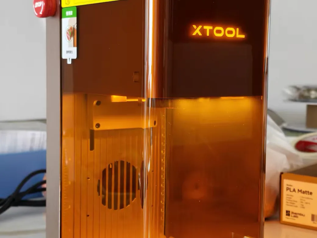 How to Choose the Best Laser Engraver/Cutter for ANY Budget 8 xTool F2 laser engraver on a desk with its glowing logo, red emergency stop button, and honeycomb bed visible