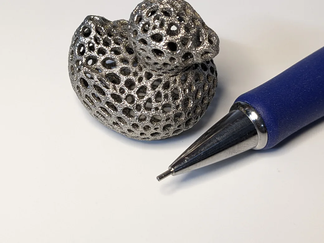 Voronoi-style rubber duck printed in 316L stainless steel on a Scrap 1 prototype, next to a mechanical pencil tip for scale